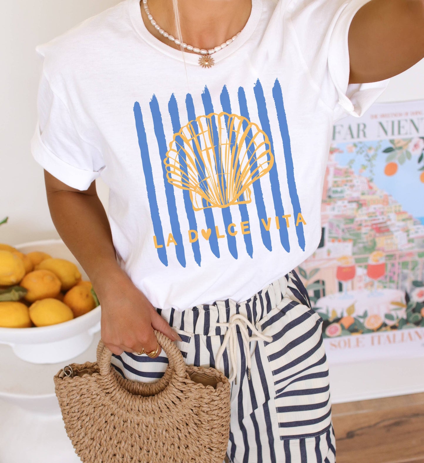 Person wearing a white t-shirt with a shell design and text, holding a woven bag in a casual setting.
