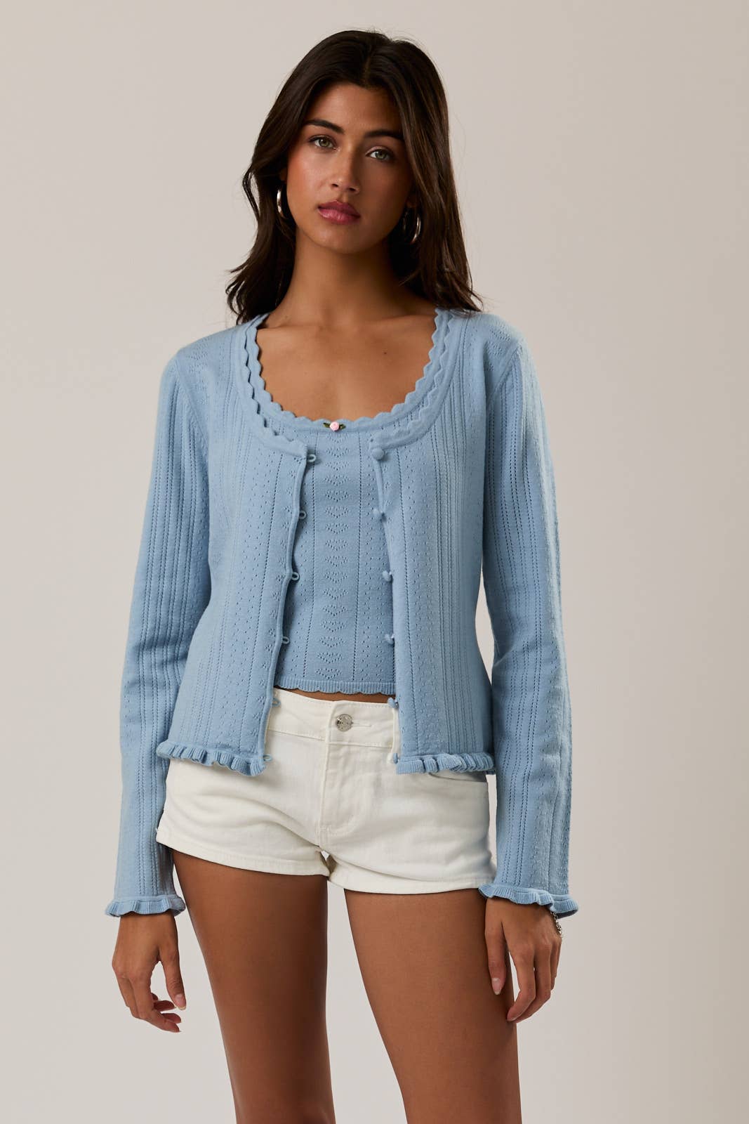 Woman wearing a light blue cardigan over a white top and shorts on a plain background