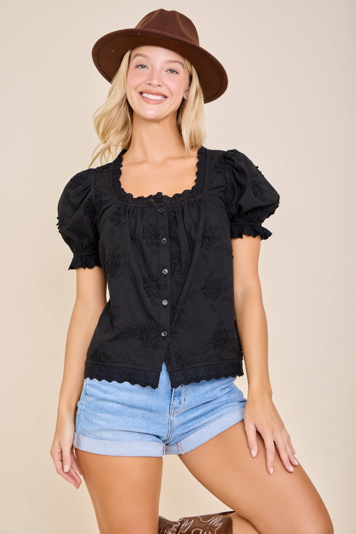 Woman wearing a black top and denim shorts with a brown hat on a beige background