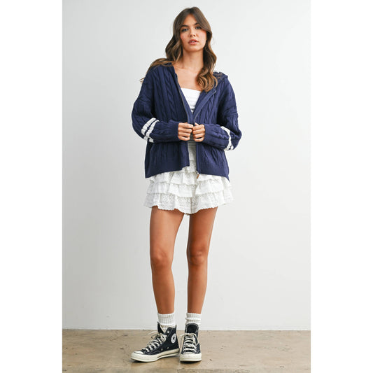 Woman wearing a blue hooded cardigan, white top, and white skirt with black sneakers on a plain background