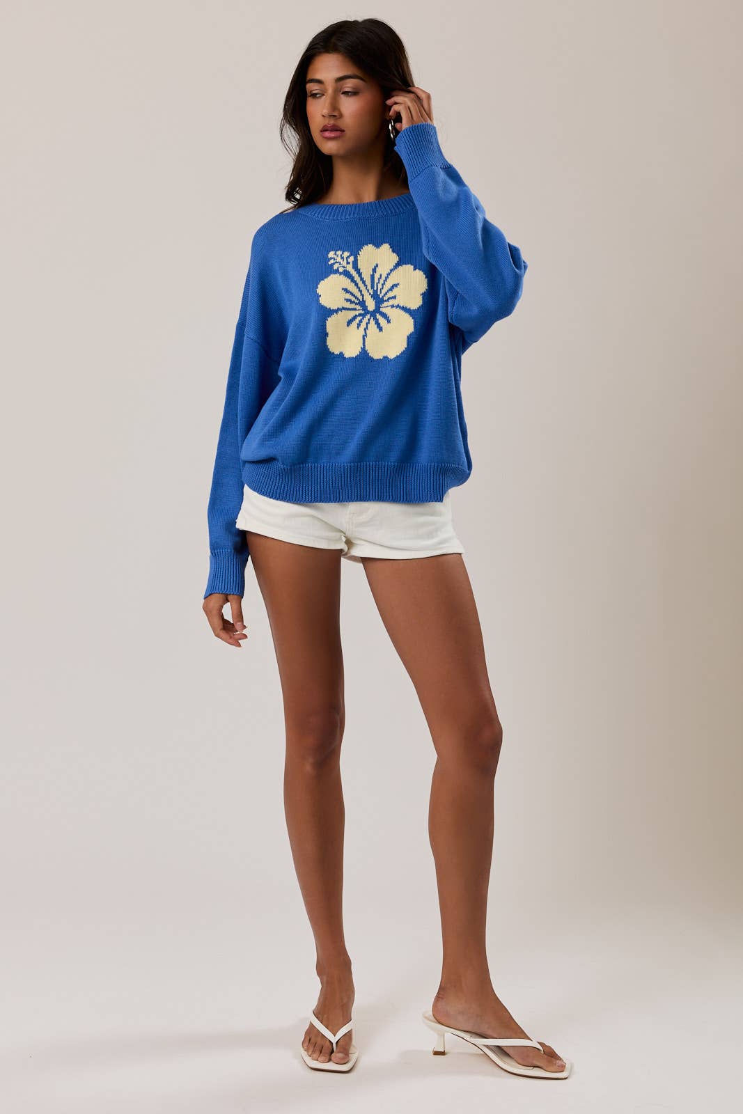 Woman wearing a blue sweater with a floral design and white shorts on a plain background