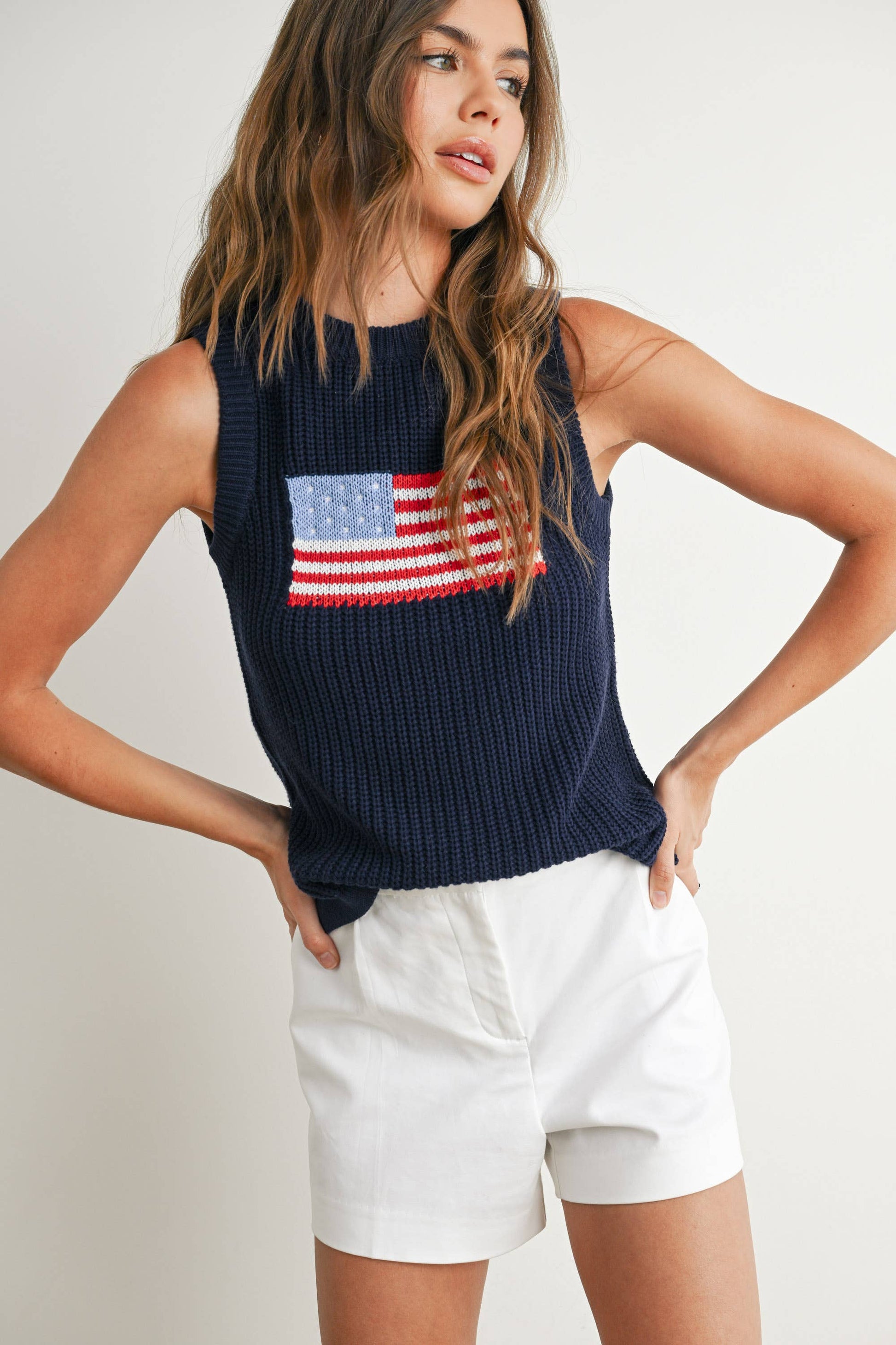 Woman wearing a navy sleeveless top with an American flag design and white shorts on a plain background