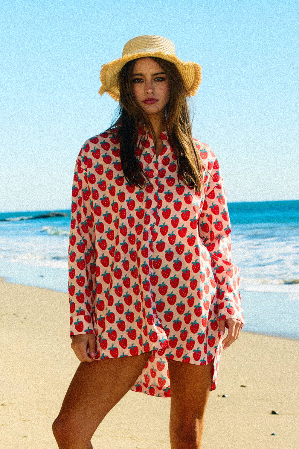 Woman wearing a strawberry patterned cover-up and straw hat on a beach.