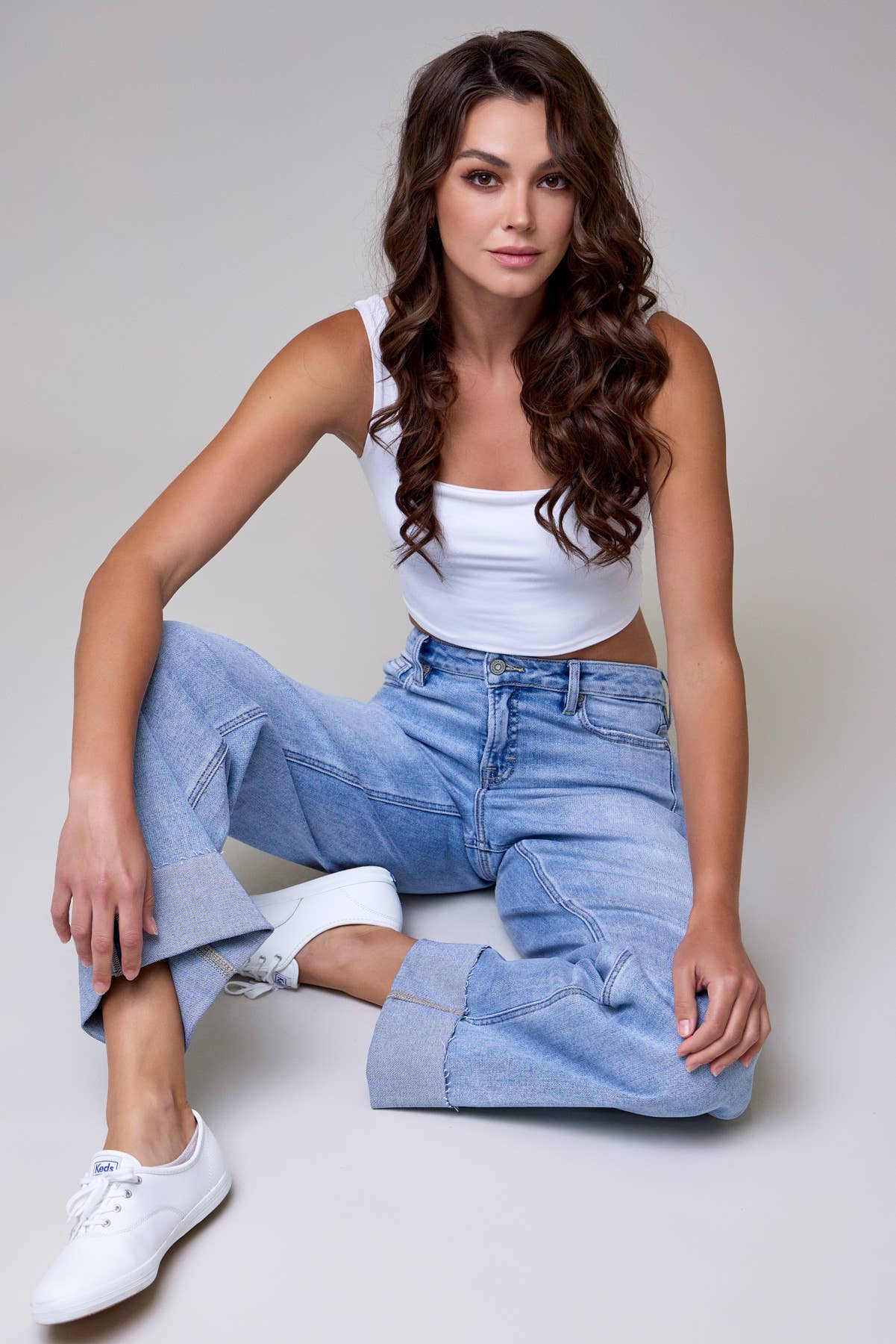 Woman wearing a white tank top and blue jeans sitting on a light gray background