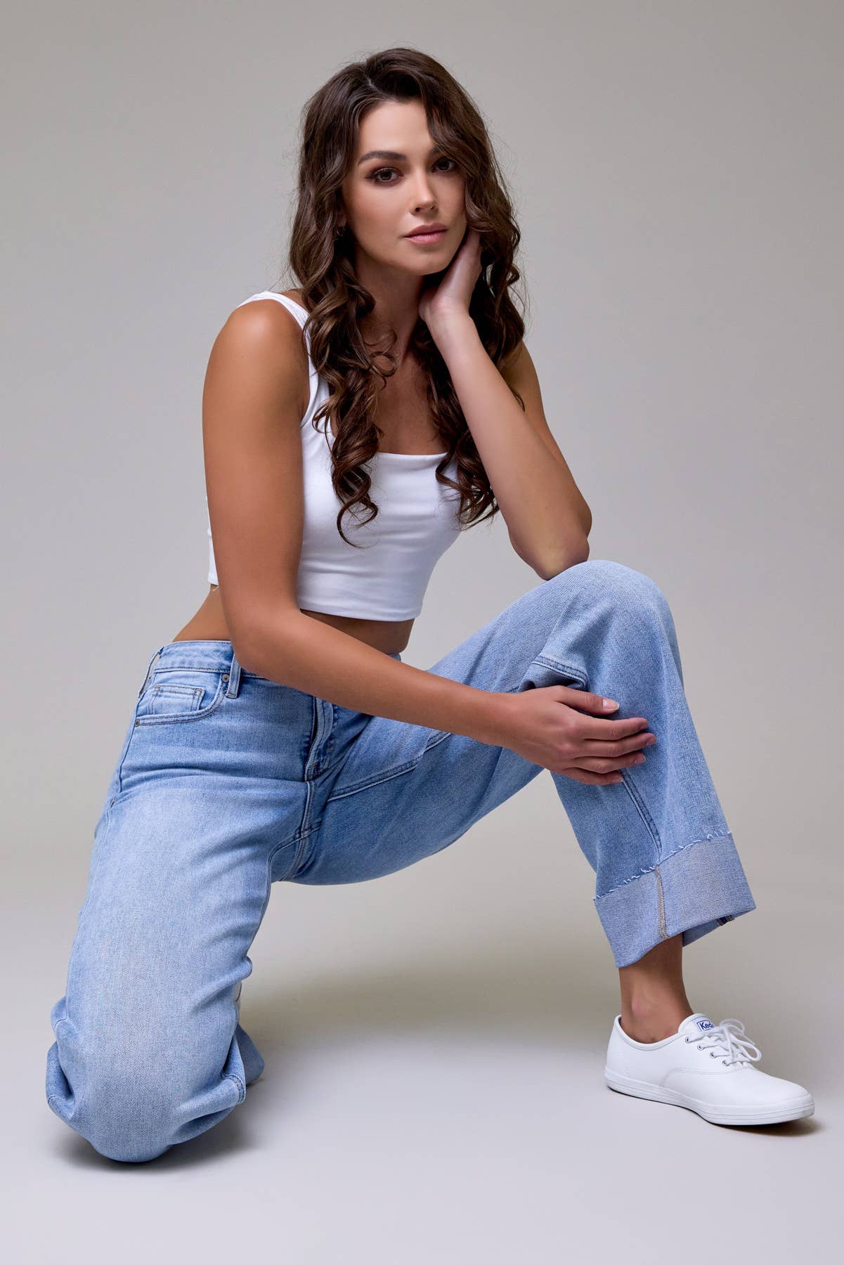 Woman wearing a white tank top and blue jeans posing against a plain background